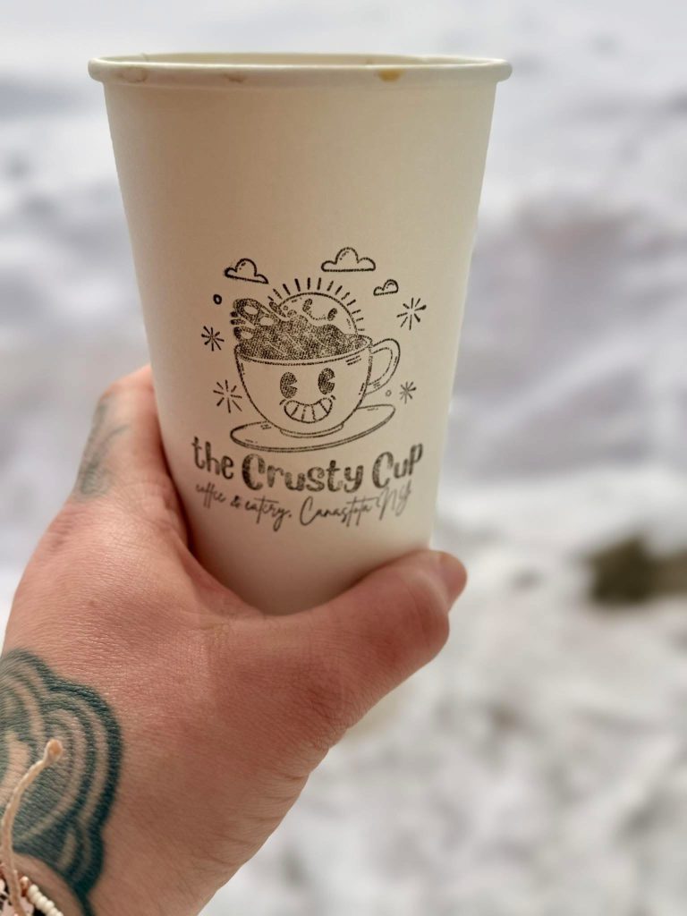 To Go Cup with Logo and left hand holding it with a tattoo in the lower left corner. Background is blurred